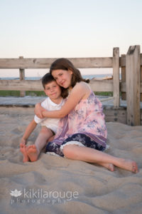 Brother and sister hugging sitting in sand by beach boardwalk