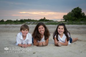 Mom with son and daughter laying in sand with sunset in background at beach
