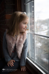 Little blonde girl in pin vest looking out widow with light smile