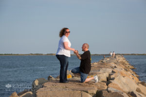 Man in black on knee on breakwater putting engagement ring on finger