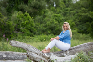 girl in white pants and blue top sitting on driftwood at beach