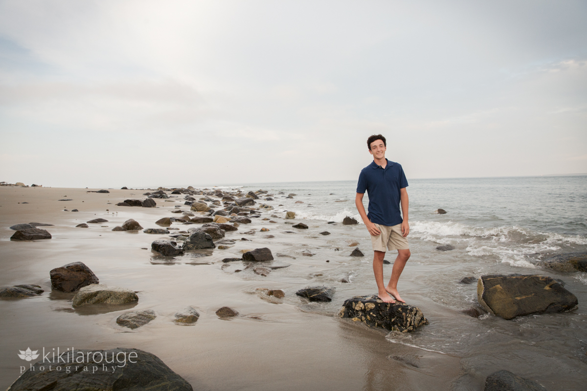 Teen boy blue shirt standing on rock at water's edge beach