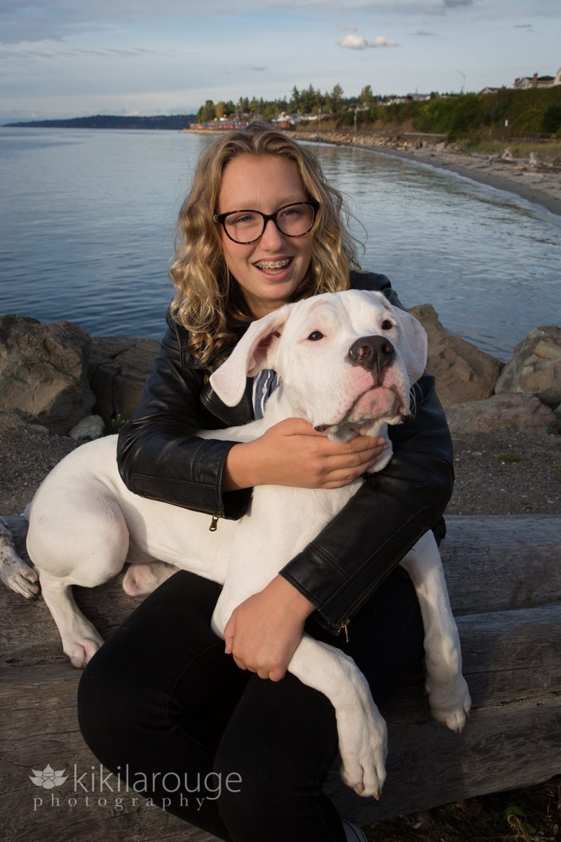 Teen girl blonde hair glasses with big white puppy on lap at beach