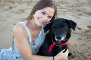 Girl with long brunette hair smiling with black dog on beach