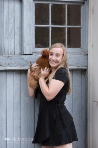 Girl in black dress by grey barn door holding a chicken
