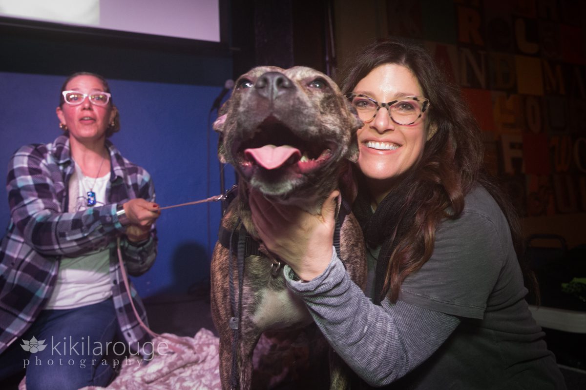 Woman in glasses smiling and holding head up of brindle pit bull mix dog at event
