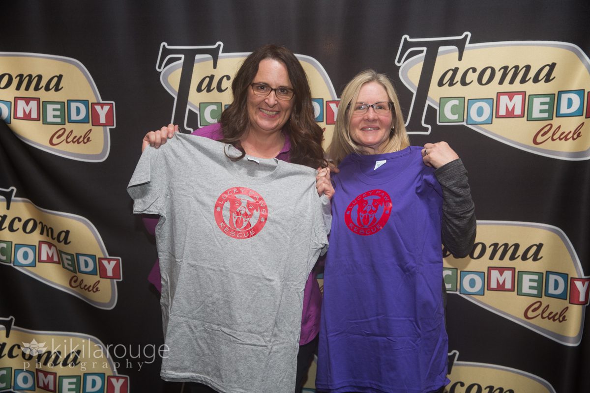 Two women in front of Tacoma Comedy Club backdrop with Lucky Paw Rescue tees