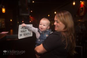 Little baby in overalls holding on to Please Wait to Be Seated Sign
