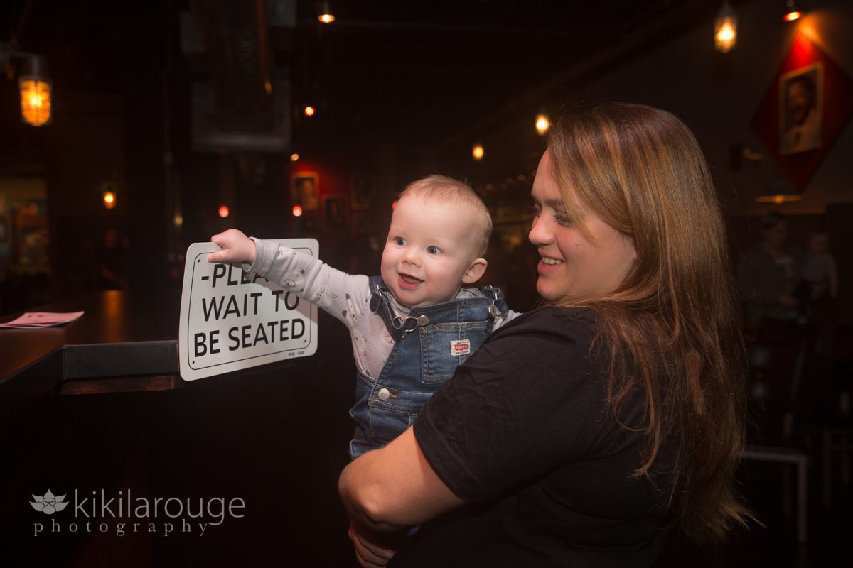 Little baby in overalls holding on to Please Wait to Be Seated Sign