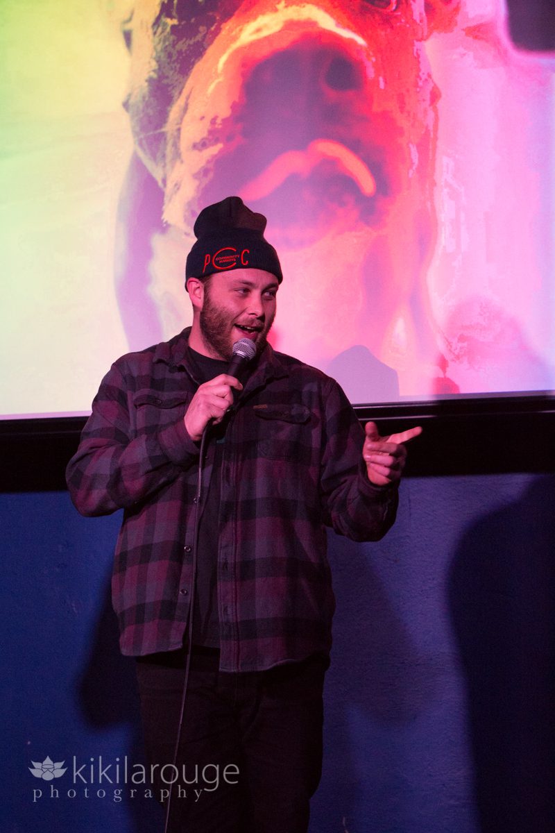 Comedian in flannel shirt and hat telling joke on stage
