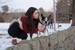 Teen girl in maroon shirt kissing Boston Terrier on stone wall with snow