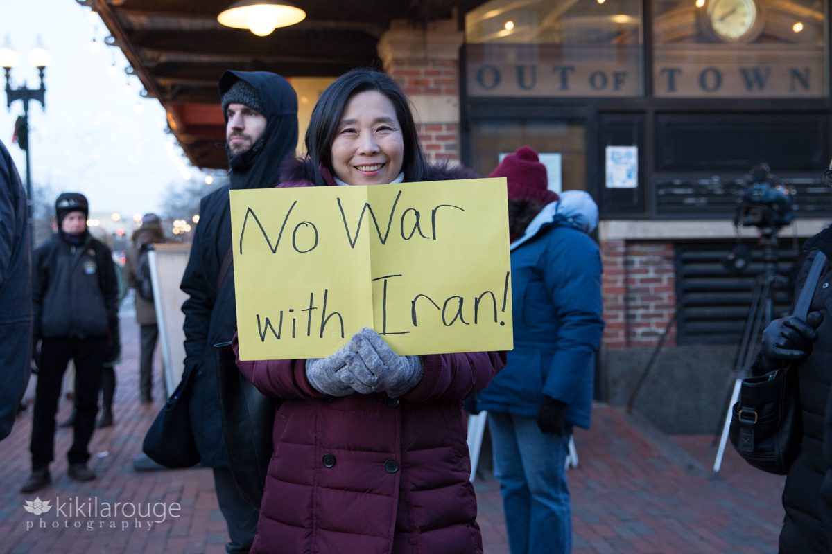 Woman in maroon winter coat at a protest with yellow sign "No War With Iran"