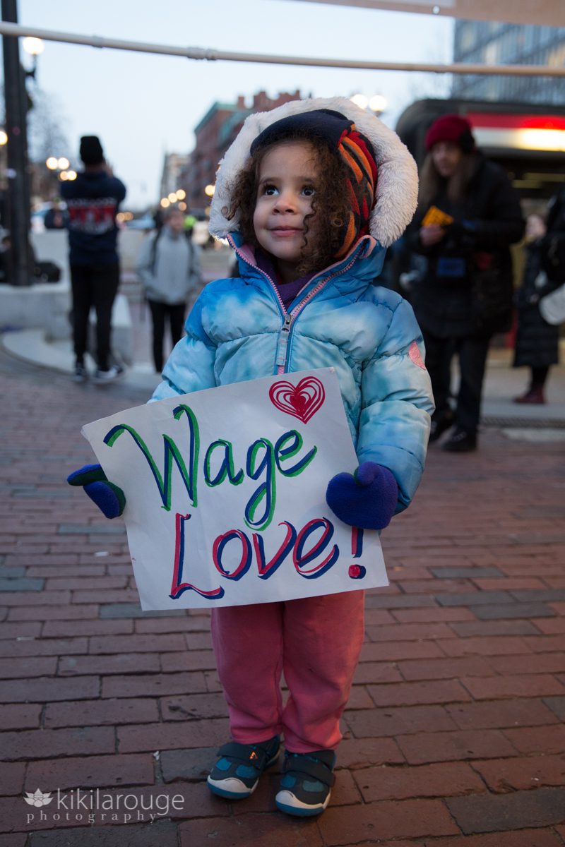 Toddler girl with curly hair in blue winter coat with a WAGE LOVE sign