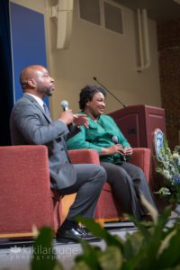 Bald man gray suit sitting on stage with a woman in green top