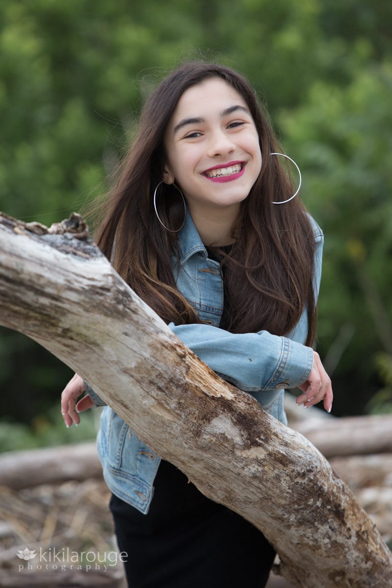 Teen girl leaning on driftwood with big smile hoop earrings and denim jacket