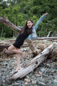 Girl with big smile waving arm holding on to beach driftwood