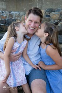 dad smiling with two daughers kissing him sitting on rock wall