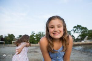 Close up wide angle portrait of young girl at beach sister running in back