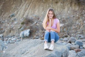 Girl in jeans and short sleeve purple top sitting on large rock at beach