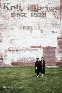 Two grads in cap and gown in front of historic mill building UMass Lowell