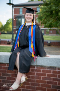 Woman sitting in cap and gown on brick wall on campus