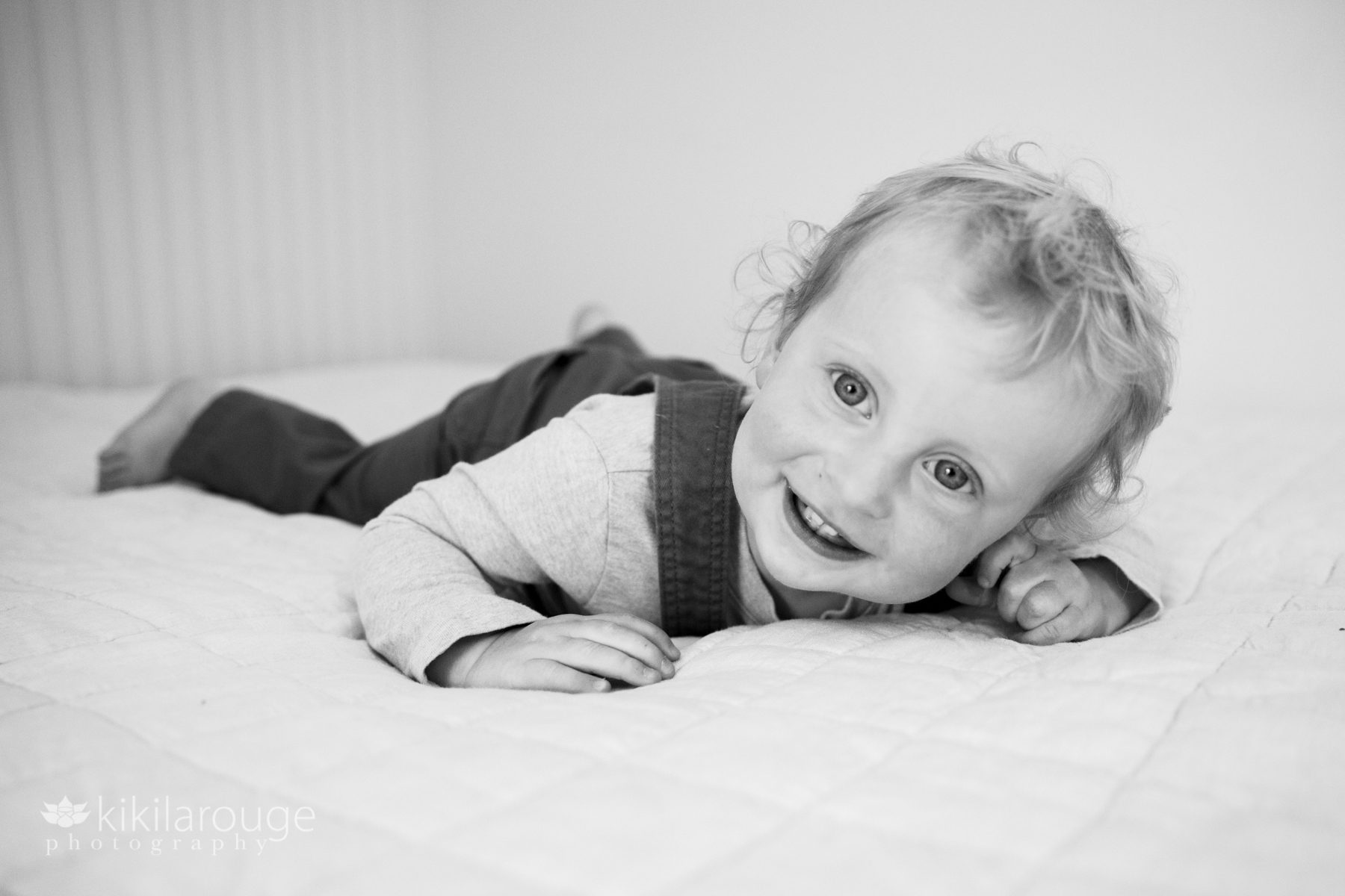 Toddler in overalls laying and smiling on bed