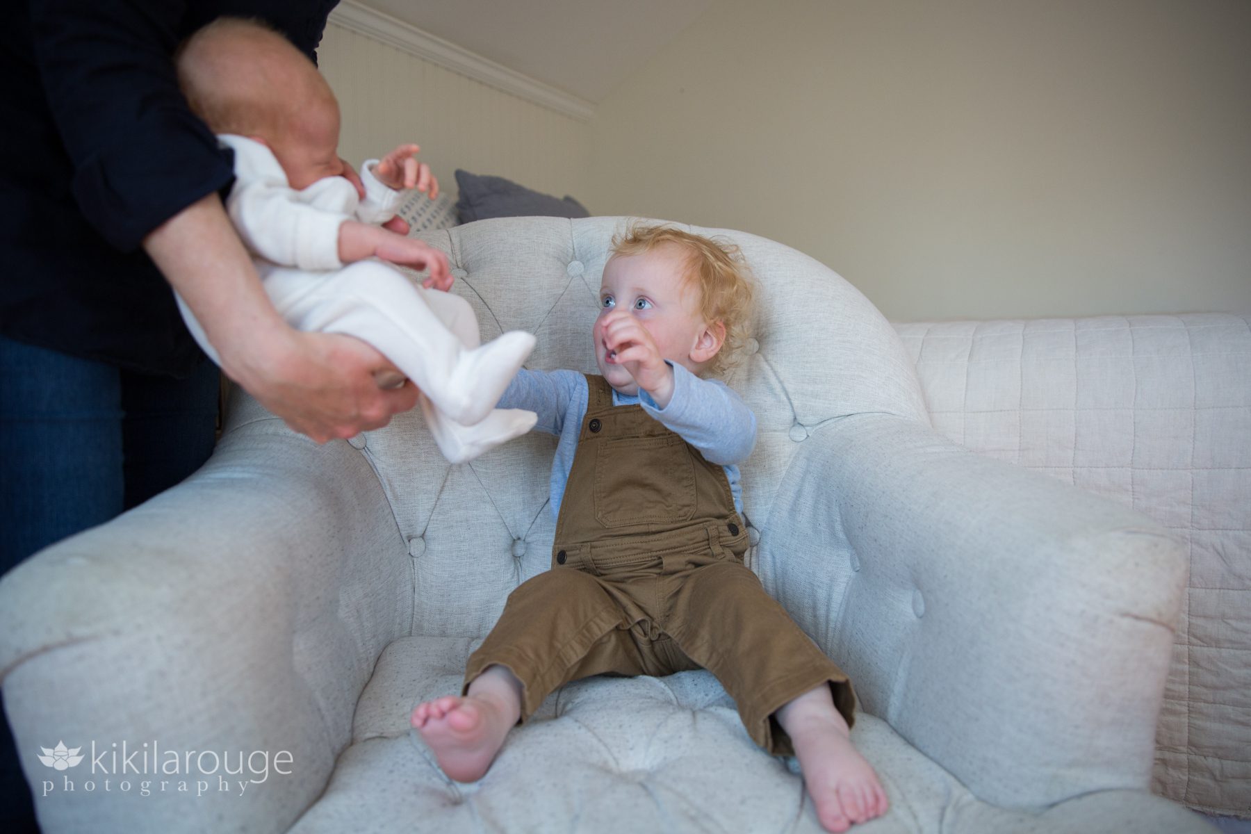 Toddler in brown overalls in chair reaching for his newborn baby sister