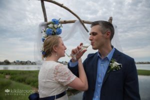 Couple sipping champagne on beach wedding day by driftwood altar