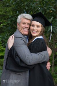 College graduate in cap and gown hugging Dad with gray blazer