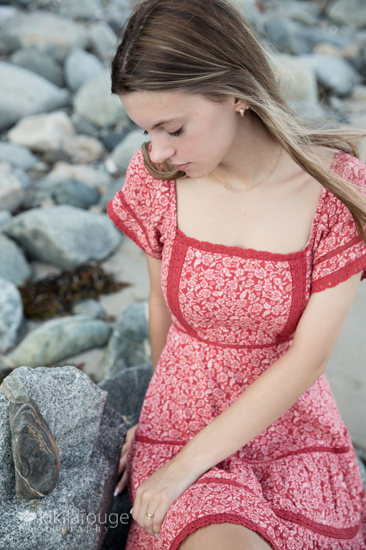 Teen girl in pretty read dress sitting on rocks at beach looking down