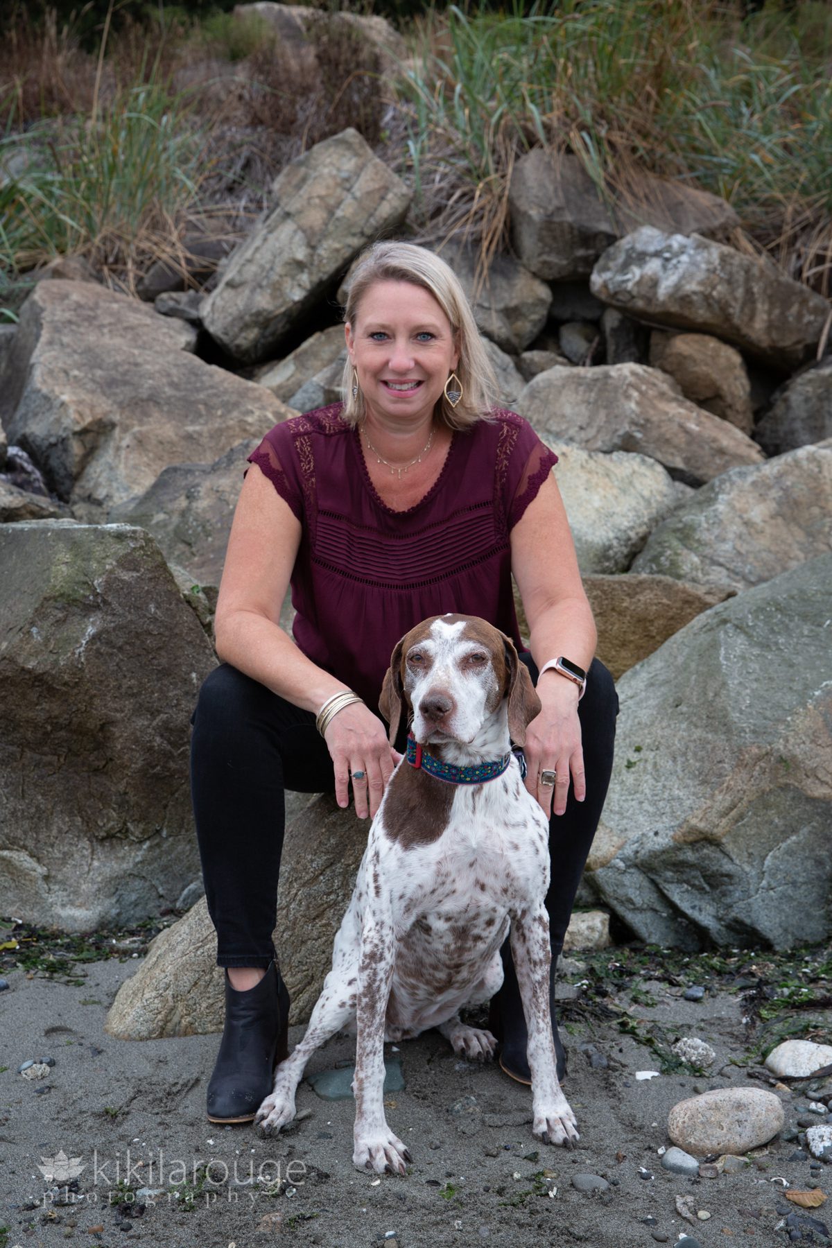 Blonde woman in maroon top with black denim with her dog at beach