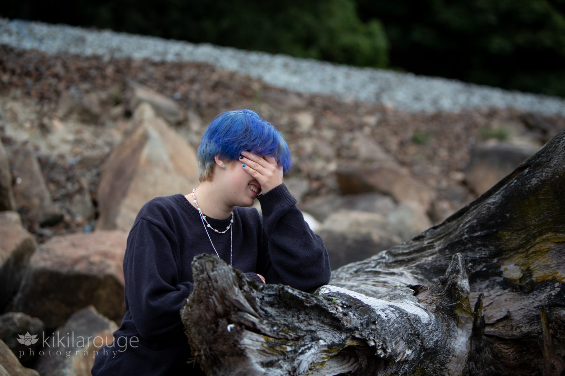 Teen with blue hair laughing into her hand leaning on large driftwood