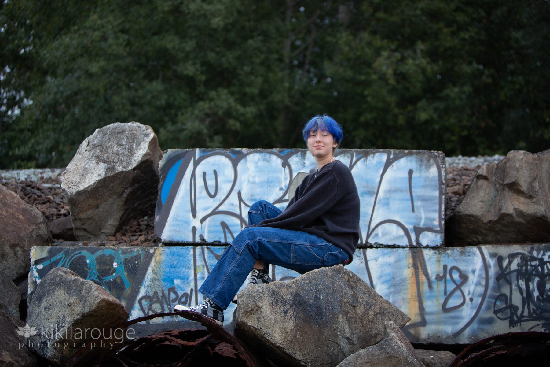 Teen with blue hair sitting on rocks in front to blue graffiti on train tracks behind