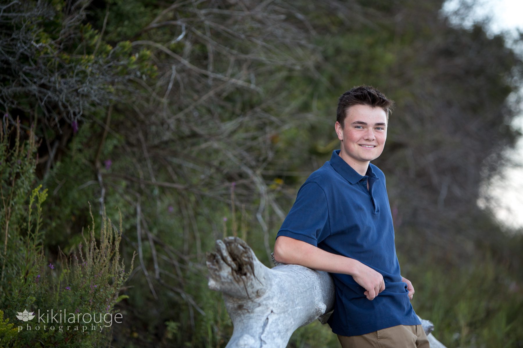 Boy in dark blue polo shirt leaning on driftwood