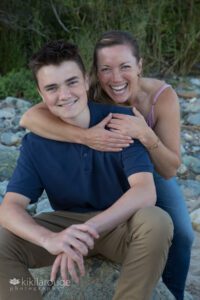 Mom hugging teen boy in blue shirt sitting on rock at beach