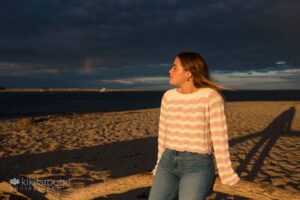 Girl sitting on driftwood looking off at sky with rainbow
