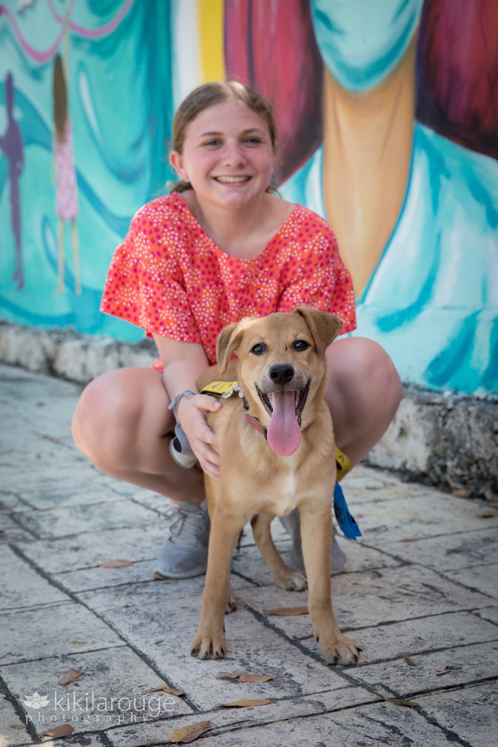 Young girl in orange floral shirt posing on sidewalk with tan puppy with tongue out
