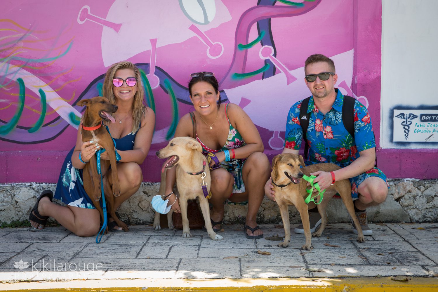 Three people in floral print by colorful purple mural in Mexico holding three rescue dogs