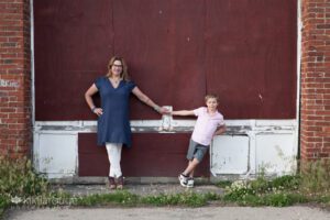 Woman in blue top and white pants holding hands with young boy in pink tip spread out together by maroon wall