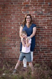 Woman in blue tunic top and white pants with young boy who is laughing by a brick wall