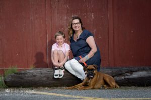 Young boy sitting on log with Mom and their boxer pup with shadows on maroon back drop newburyport ma
