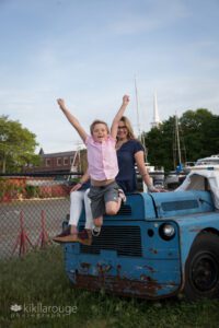 Young boy jumping off blue tractor in NBPT boat yard with Mom in background