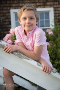 Blonde boy in pink top leaning on white stair post with pink flowers in bloom in back ground