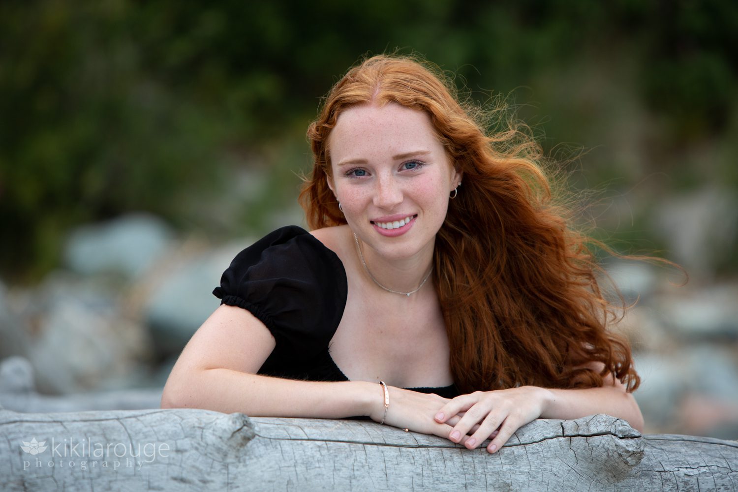 Redheaded girl smiling leaning on driftwood at beach with black top