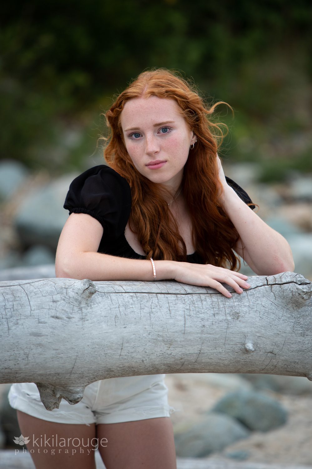 Girl with red hair and black top white shorts leaning on beach driftwood serious face