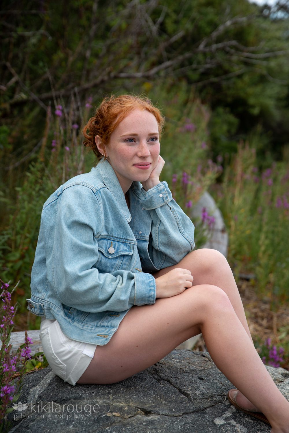 Redhead with denim jacket and white short silting on rock with beach flowers