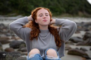 Girl with hands in hair eyes closed looking up in ripped jeans sitting at rocky beach
