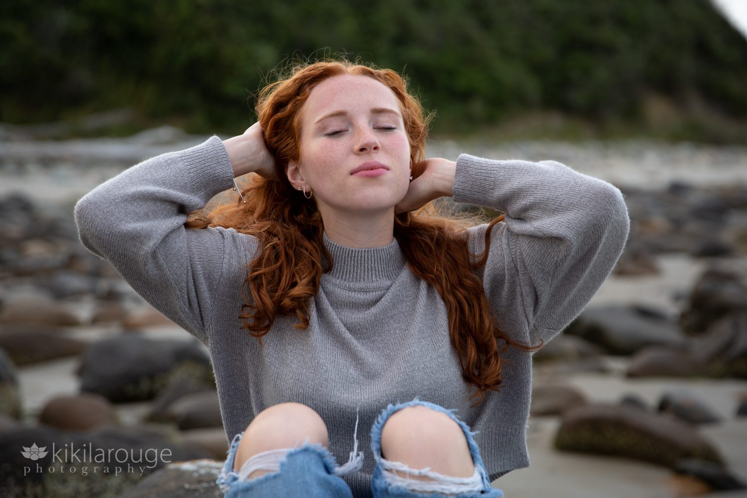 Girl with hands in hair eyes closed looking up in ripped jeans sitting at rocky beach