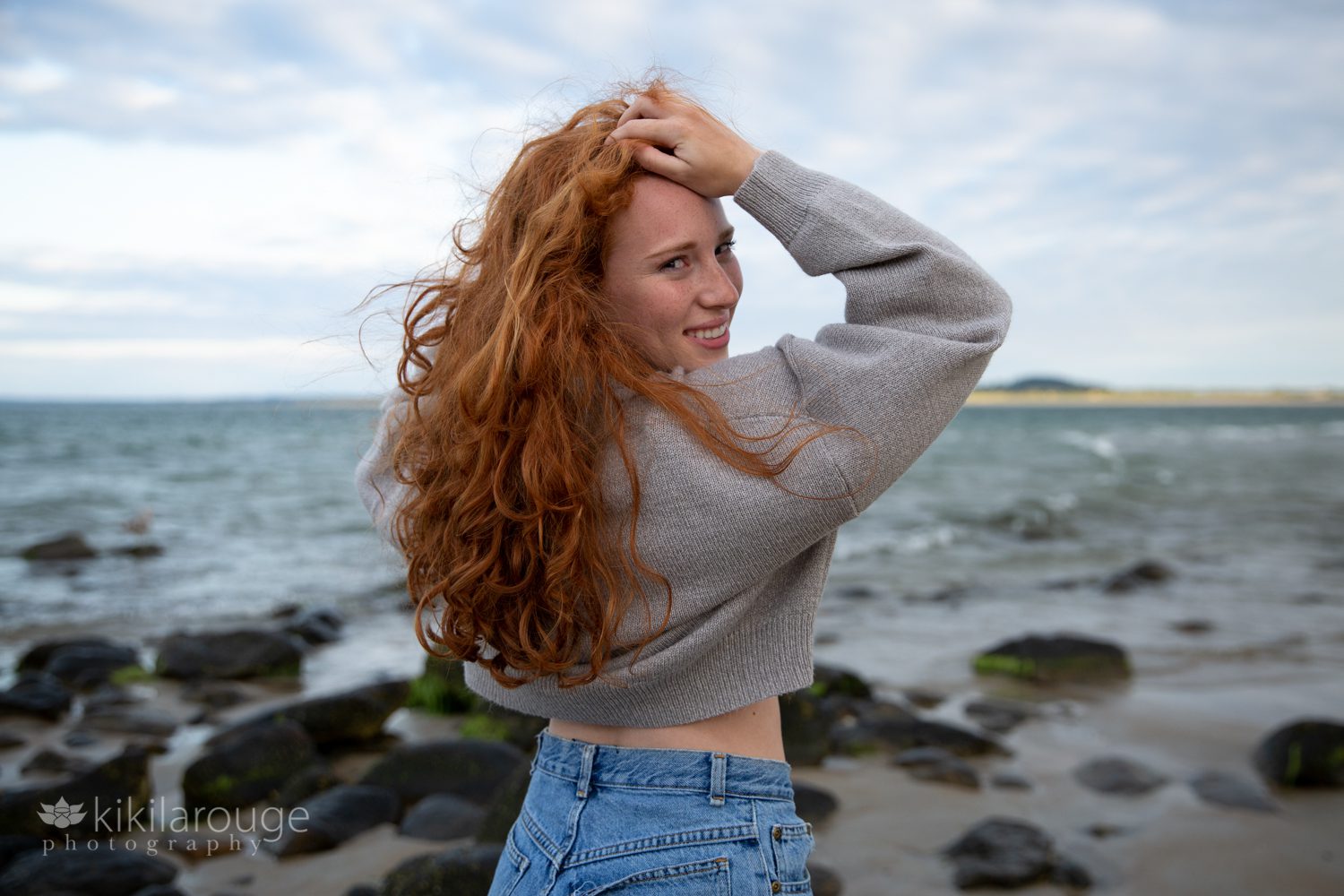 Redhead girl with long hair looking over shoulder at beach smiling with grey sweater on