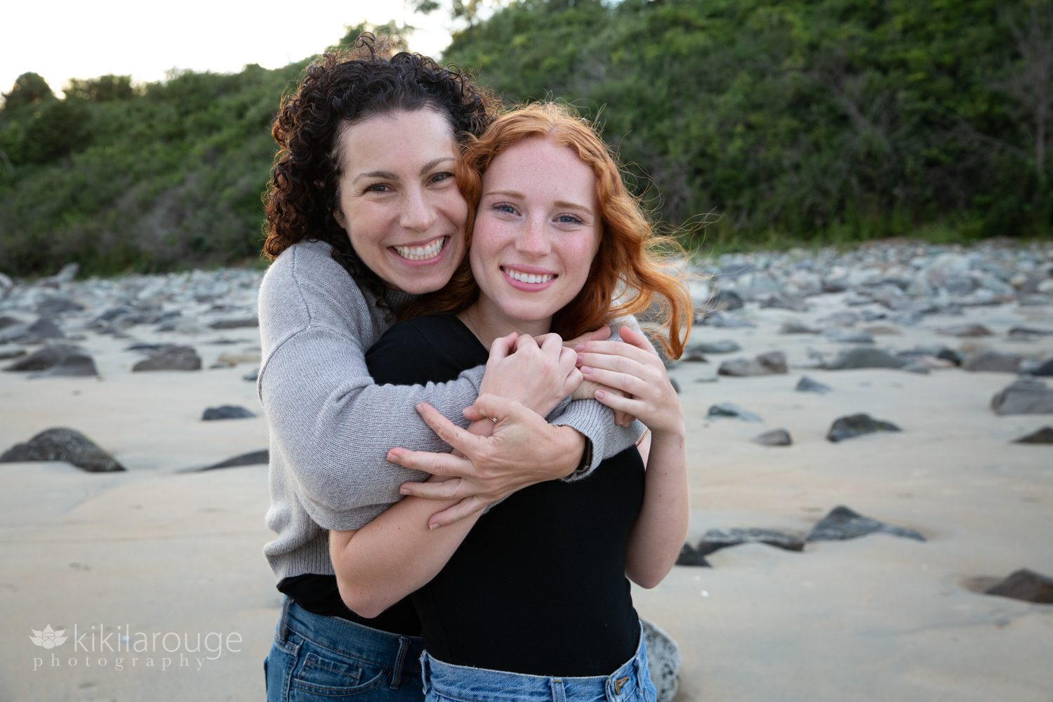 Portrait of mom and teen daughter hugging at beach with rocks in background